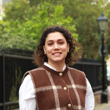 Elifcan has shoulder-length, curly dark hair. She is wearing a white shirt and a brown vest with a large white check. She is standing in front of a park fence, smiling. 