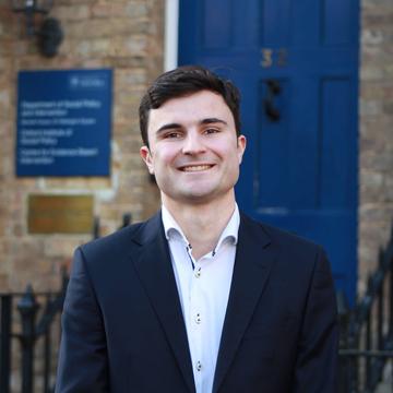 Isaac Mellis-Glynn Stands in front of the DSPI building. He has dark brown hair and wears a white shirt and navy blazer.