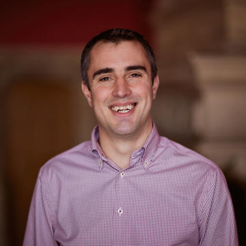 Peter Burr is wearing a purple shirt, with short dark hair and is smiling at the camera.