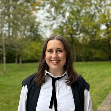 Quinn Sanderson wears an academic gown and is standing in front of a green lawn.  