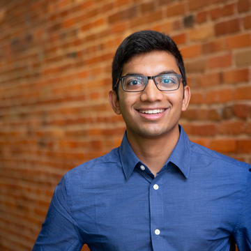 Shreyas wears glasses, has short black hair, and is wearing a blue shirt.