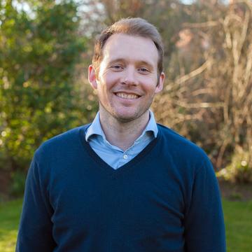 Alex Sutherland has short fair hair and is wearing a light blue shirt with a navy jumper. He is standing outside in a garden, smiling at the camera. 