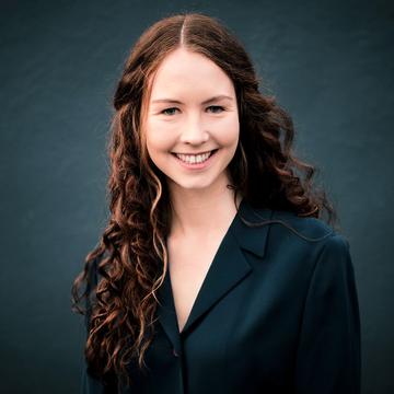 Tamsin Greene Barker is smiling at the camera, with long, dark, shoulder-length curly hair, wearing a dark jacket.