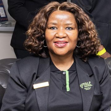 Tlale has curly, shoulder-length brown hair. She is wearing a black polo shirt and blazer, smiling at the camera.