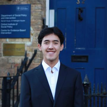 Xin Law-Gallagher stands, smiling, in front of the DSPI blue door. He wears a white shirt and dark blazer. He has short cropped dark hair. 