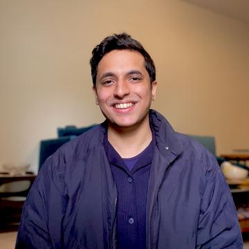 Ahsan smiles broadly at the camera. He has short brown hair and is wearing a navy blue knitted shirt and jacket. 