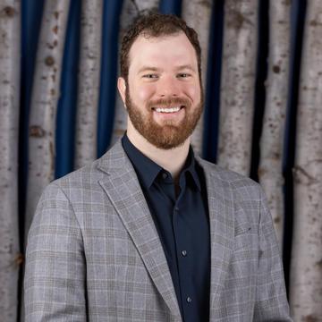 Alex Coccia smiles, standing in a grey suit jacket and dark-blue shirt 