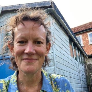 Amanda has curly brown hair which is partially tied back. She is wearing a patterned green and blue blouse, and sat outside in front of a small wooden structure.