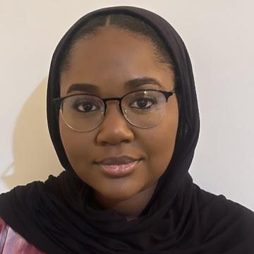A black hijab and glasses are worn by Amina Hamza, who is looking at the camera directly.