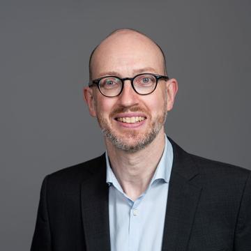 Arne Kuehl is wearing a suit and glasses smiling