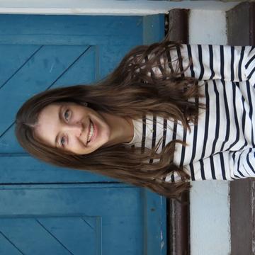 Bridget Steele has long brown hair and is wearing a black and white striped shirt. She is sitting on some outdoor steps in front of a blue door and smiling.