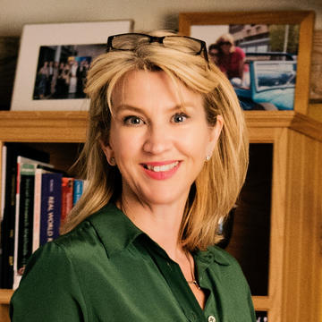 Caroline Greenhalgh wears a dark green shirt, has shoulder-length blonde hair and stands in front of a bookshelf. 