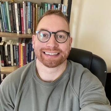 Dr Michael Franner is smiling at the camera , wearing glasses and a grey top with red hair and a beard, sitting in front of a bookcase.      