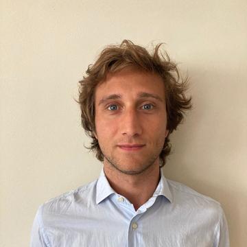 Enrico is standing in front of a wall looking at the camera. He has fair hair and is wearing a light shirt 