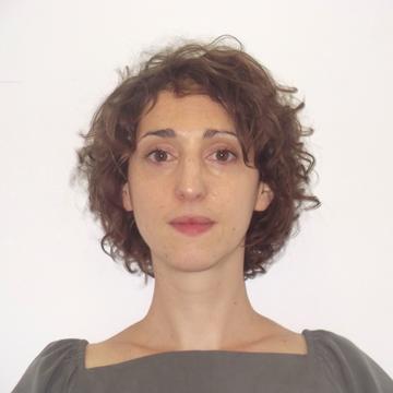 A headshot of Ilka Vari-Lavoisier. She has short curly brown hair. She is standing face-on to the camera, wearing a square-necked grey top with puffed sleeves.