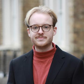 Hendrik Schäfer has blonde hair and wears glasses. He is standing in front of the DSPI building in a red polo-neck top and blazer.