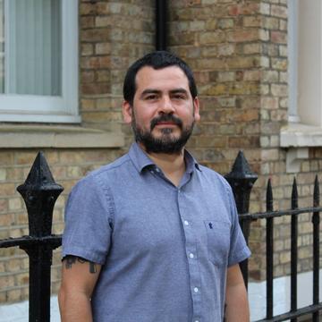 Dr Munoz has short black hair and black facial hair. He is wearing a short-sleeved blue shirt and standing outside a brick building. 