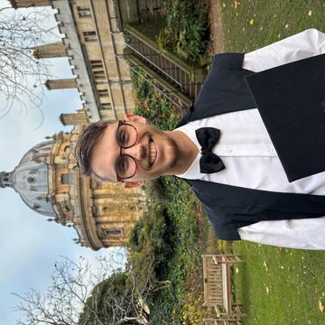 James Ward is wearing his graduation down with a white shirt, bow tie and glasses sliming at the camera.