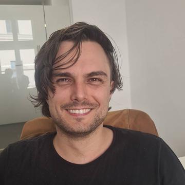 Jim Schmeckenbecher is smiling, sitting in an office. He has dark brown hair and is wearing a dark-coloured tee-shirt.