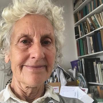 Dr Peretz has short curly white hair. She is sat in front of a large bookcase, smiling. 