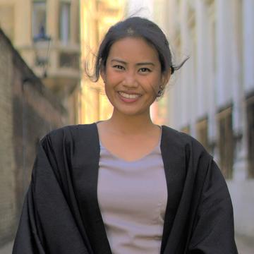 A smiling Marianne stands in a narrow street wearing a black graduation gown.