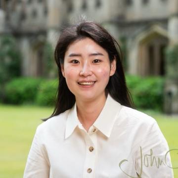 Seonwoo, with shoulder-length black hair, wearing a white shirt, stands on college grounds in front of a building, smiling at the camera.