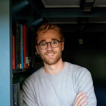 Robert Hill has short fair hair and is wearing round tortoiseshell glasses and a grey jumper. He is leaning against a bookshelf.