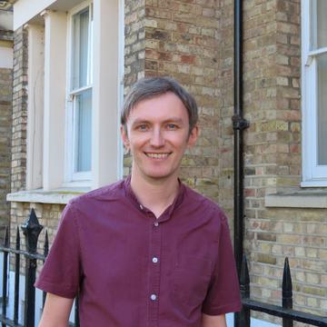 Robin has short fair hair and is wearing a short-sleeved burgundy shirt. He is standing outside in front of some light-brown brick buildings.