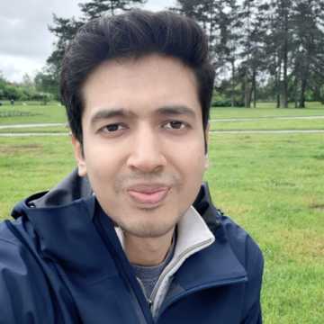 Mobarak is looking directly at the camera, wearing a dark blue top with short brown hair standing in a field.