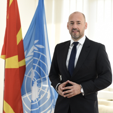 Mr. Benjamin Perks is wearing  a suit and tie standing in front of flags including at UN flag.