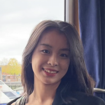 Yaxin has long dark hair. She is standing in front of a window and smiling. 