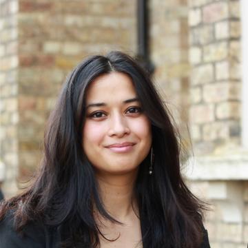 Srishti Rongpipi has long brown hair and is smiling in front of a building.