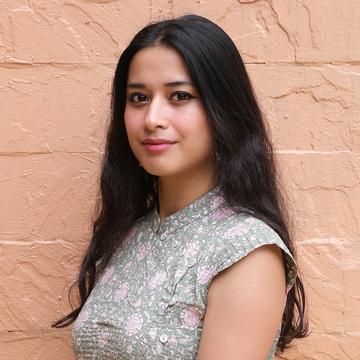 Srishti Rongpipi has long black hair, wearing a sage patterned top and is standing in front of a pink wall