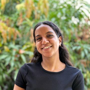 Tejashwari has long dark hair. She is wearing a black short sleeved t-shirt. She is standing in front of some foliage, smiling. 
