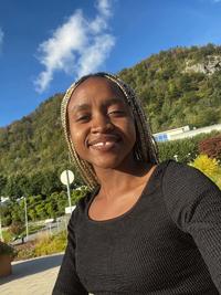 Diana Mwala is smiling in front of a blue sky. Her hair is braided and there is a tree-lined hill in the background. 