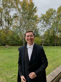 George Rappa is smiling in front of a patch of grass. he is wearing an academic gown, with dark suit and white tie. He also wears glasses and is carrying a mortar board. 