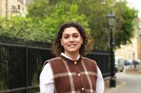 Elifcan has shoulder-length, curly dark hair. She is wearing a white shirt and a brown vest with a large white check. She is standing in front of a park fence, smiling. 
