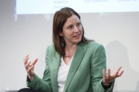 As Silja Häusermann sits on a stage wearing a green suit, she appears to be in a discussion and expressing herself with her hands. 