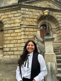 Lhori Webster is wearing academic dress and stands in front of a sandstone building in Oxford. 
