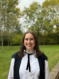 Quinn Sanderson wears an academic gown and is standing in front of a green lawn.  