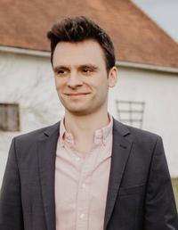 Michael Ganslmeier a DSPI doctoral researcher is smiling while wearing a dark grey suit and a salmon shirt.