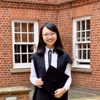 Yiwei Zhou stands in a courtyard wearing academic dress. She is smiling, holding a mortar board.