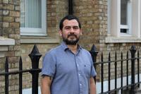 Dr Munoz has short black hair and black facial hair. He is wearing a short-sleeved blue shirt and standing outside a brick building. 
