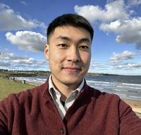 Man with short dark hair, wearing a jumper and standing near the sea
