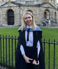Woman with blond hair, wearing university gown standing by a metal rail with University building in background