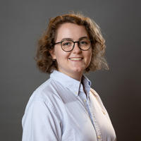 Leah Ennis waers a lab coat and glasses and is smiling in front of a grey background