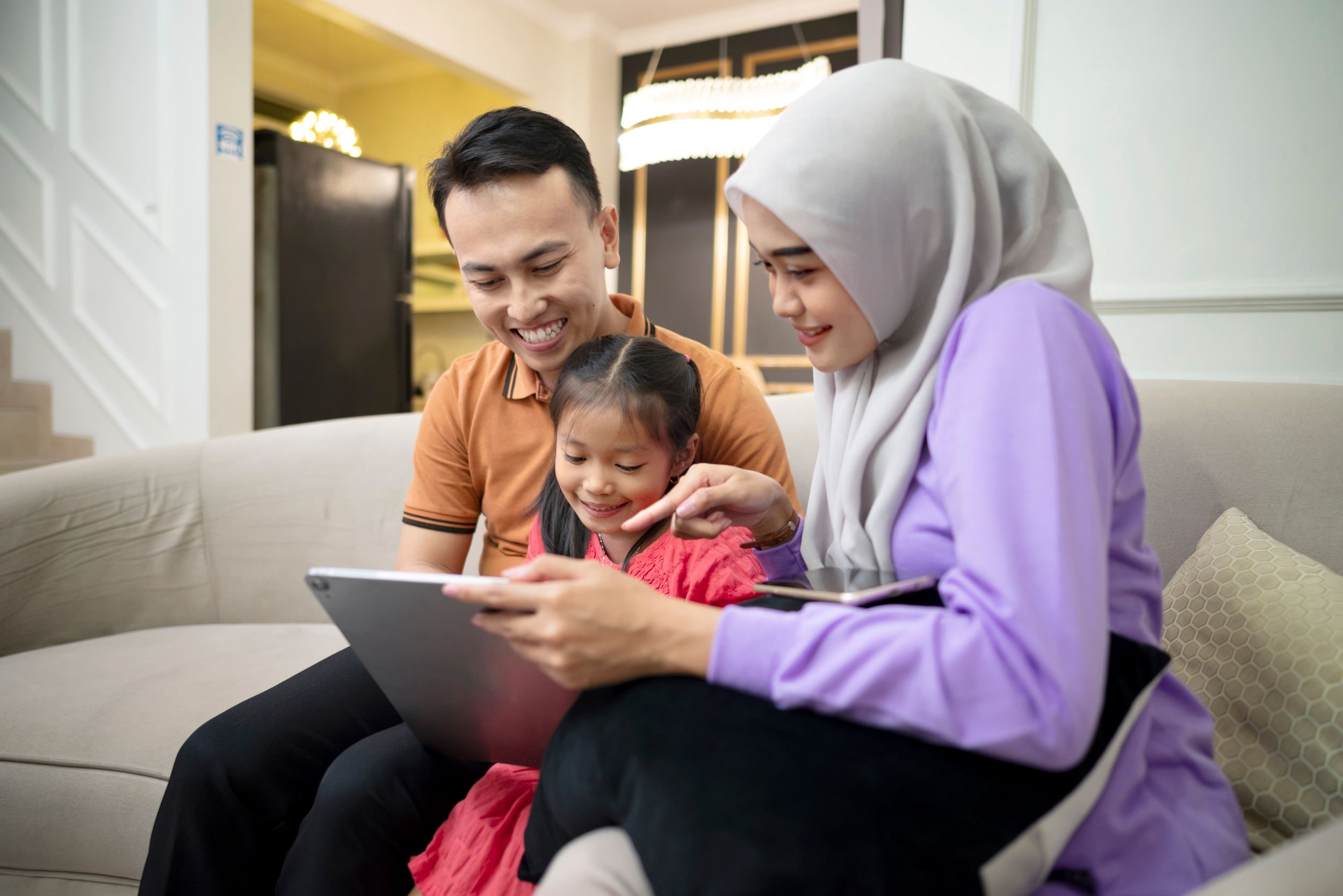 A mother and father relaxing with their young daughter, holding a digital tablet and sitting on a sofa