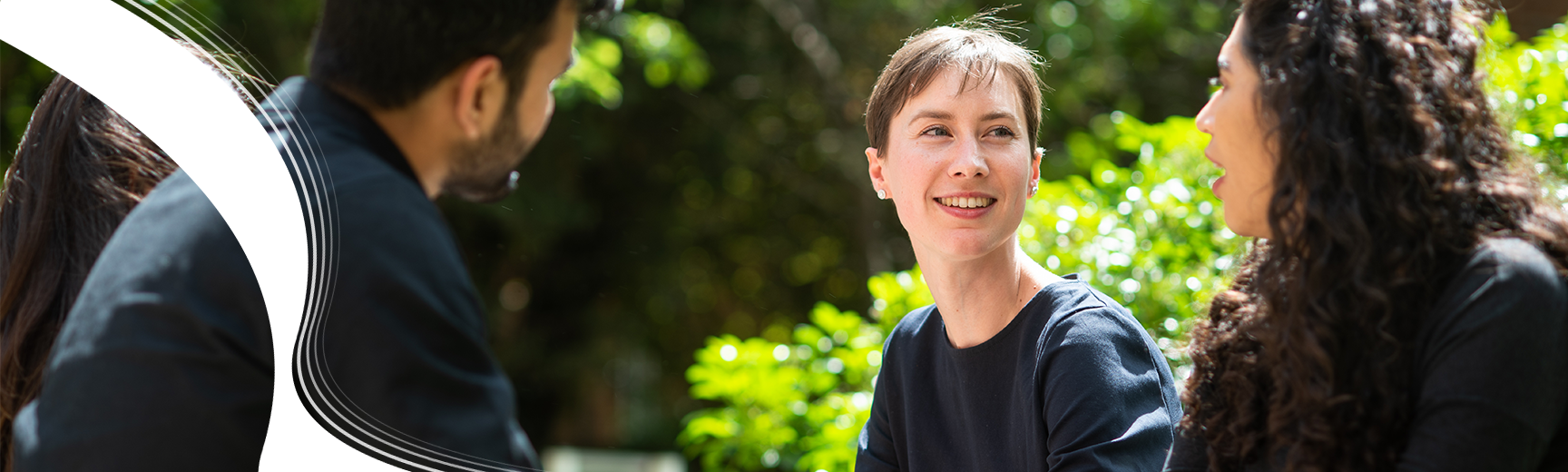 Three students sat outside in the sunshine, chatting. One student with short brown hair and a navy t-shirt is looking to her friend, smiling.