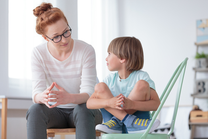 A young boy sits cross-legged on a chair, looking up at an adult woman sat next him.