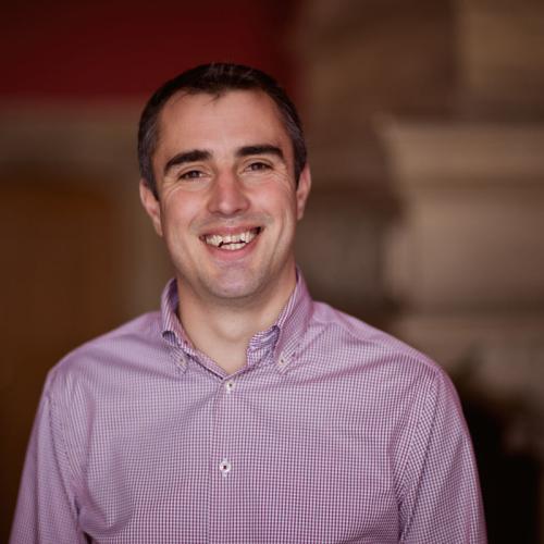 Peter Burr is wearing a purple shirt, with short dark hair and is smiling at the camera.